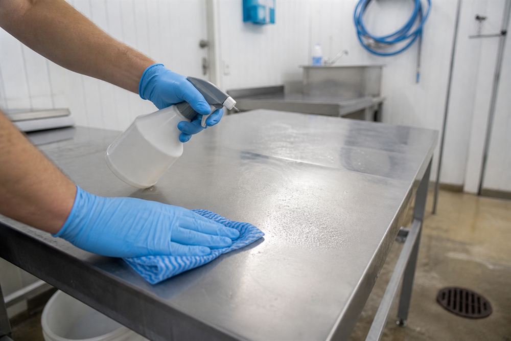Gloved hands sanitizing a stainless steel preparation surface in a bright, orderly space